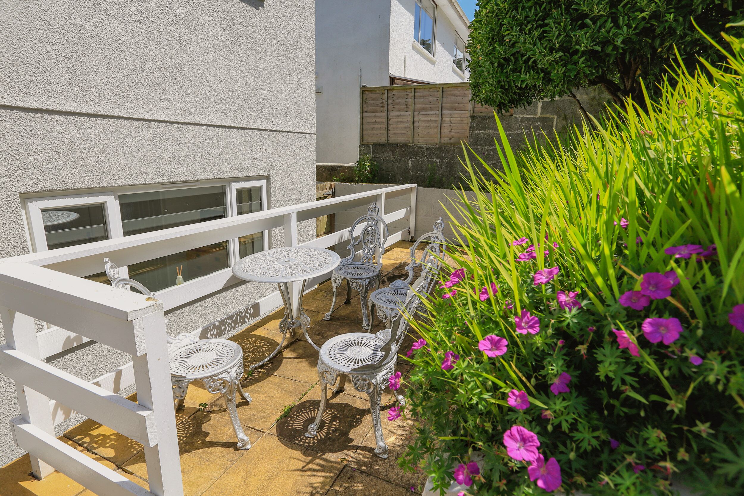 Une terrasse extérieure avec des chaises et des tables en métal blanc, entourée d’une clôture blanche et de plantes en fleurs.