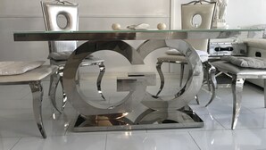 Un comedor moderno con una mesa de vidrio, sillas de cromo y una gran pieza central metálica con el logotipo 'GG'.