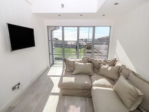 A modern living room with a large sectional sofa, a flat-screen TV mounted on the wall, and a large window offering a view of the outdoors. - Angorfa - Trearddur Bay (Holyhead)