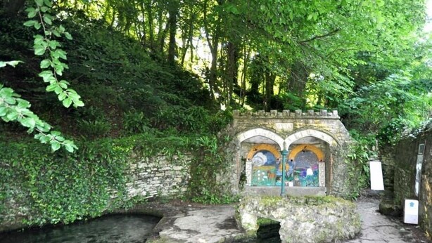 Edifici de pedra amb finestres d'arc, un petit canal d'aigua i una paret de pedra coberta de vegetació.