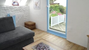Una habitación acogedora con un sofá gris, una mesita de madera y una pared blanca. Se aprecia una ventana con vistas a un jardín y una cerca blanca.