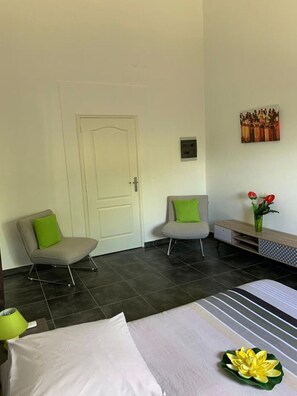 Una habitación de hotel con una cama, dos sillas con cojines verdes, una mesita con un florero y una puerta.