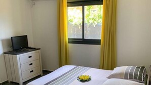 Une chambre à coucher avec un lit, une table de chevet avec une lampe, un téléviseur posé sur un meuble et une fenêtre avec des rideaux.