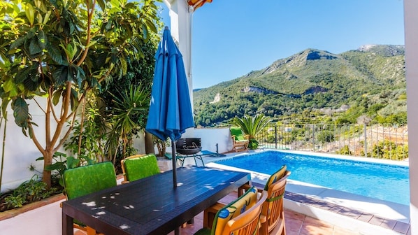 A poolside area with a table and chairs, a barbecue grill, and a view of a mountainous landscape.