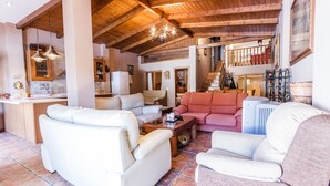 A spacious living area with a high ceiling and exposed wooden beams, featuring a kitchen area, a fireplace, and a staircase leading to an upper level. - Monte Verde Ojén by Ruralidays (Málaga)