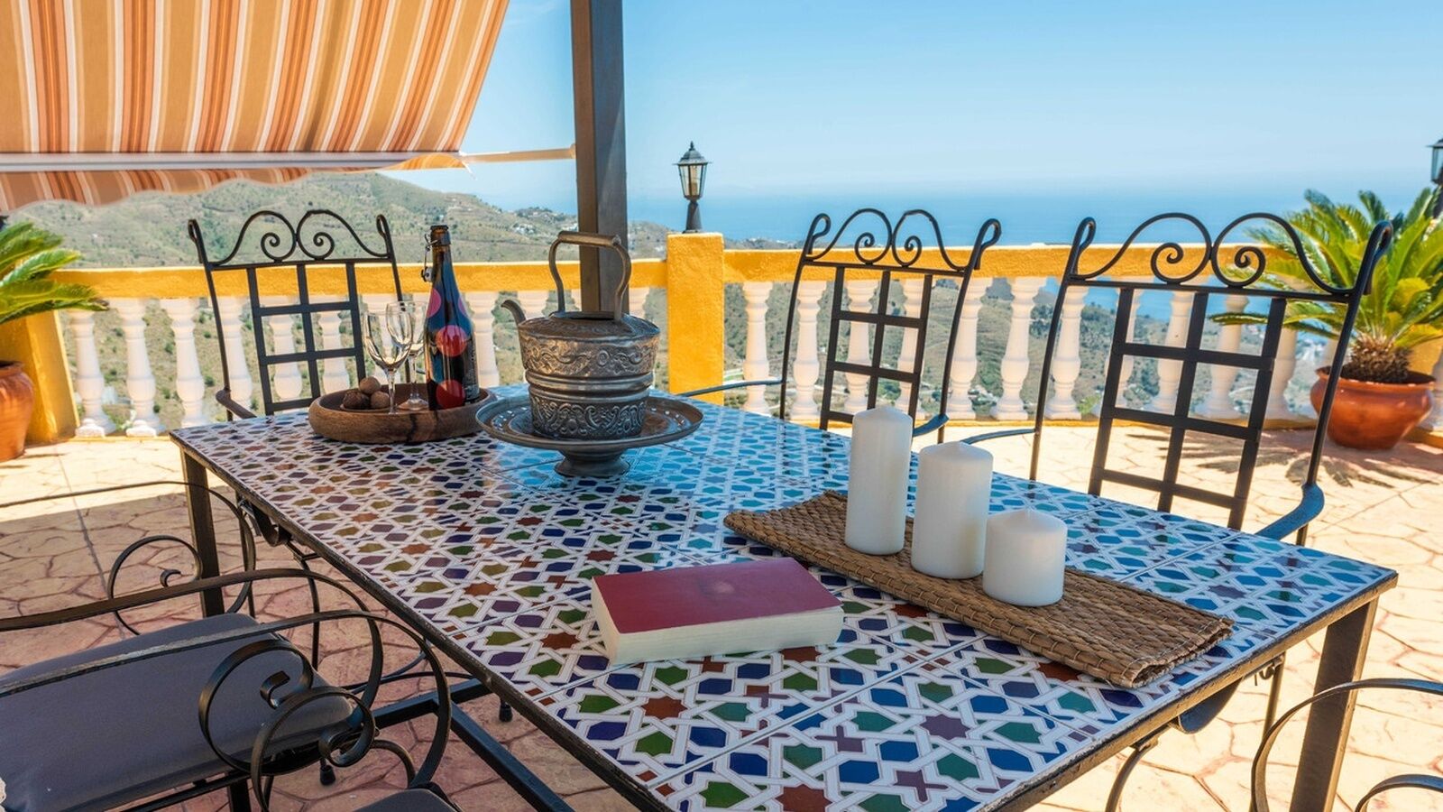 A table with a patterned tablecloth, a book, candles, a bottle, and a decorative pot on a balcony with a view of the sea.