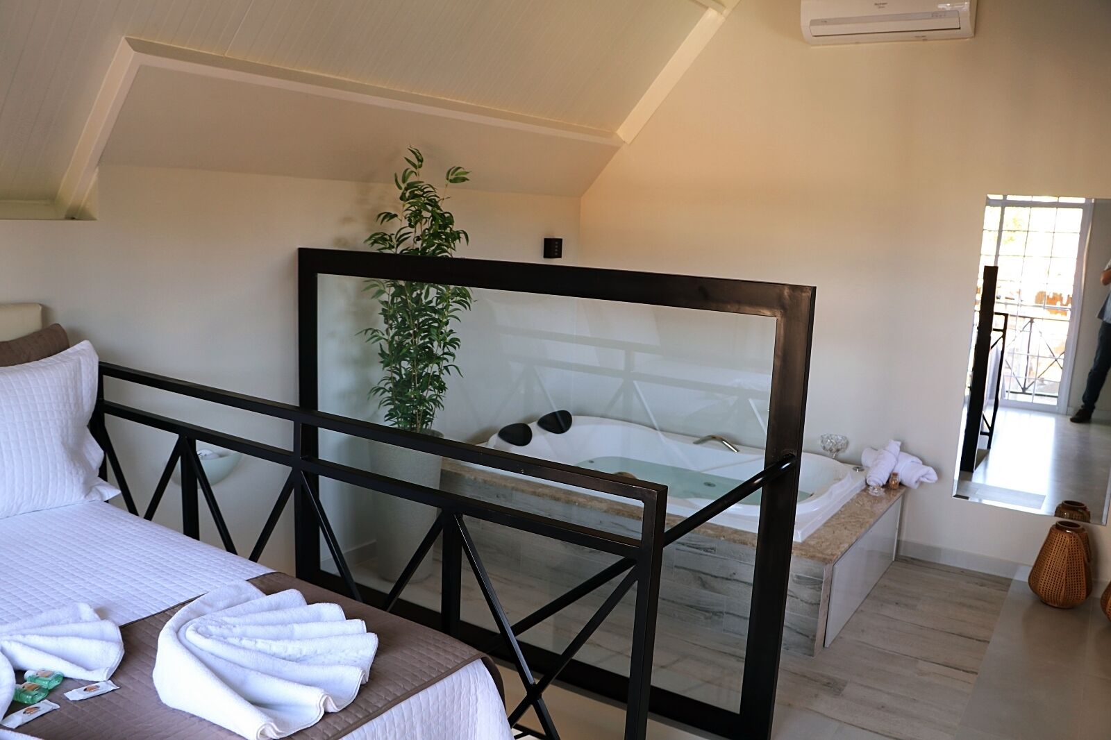 Un dormitorio con cama, mampara de vidrio, planta en maceta, bañera y baño con espejo y toallas.