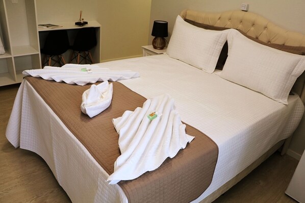 Una cama bien tendida con una colcha marrón y ropa de cama blanca, una mesita de noche con una lámpara, y una silla al fondo.