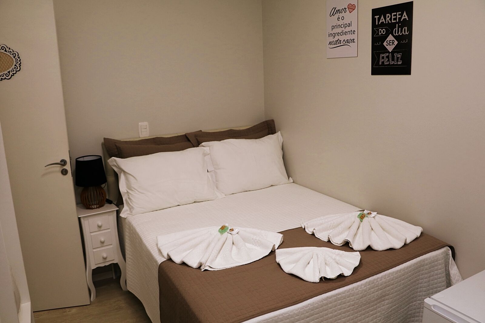 Cama bem arrumada com roupa de cama branca e marrom, mesa de cabeceira com abajur, e duas decorações de parede.