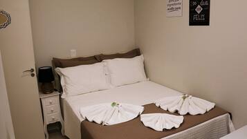 Cama bem arrumada com roupa de cama branca e marrom, mesa de cabeceira com abajur, e duas decorações de parede.