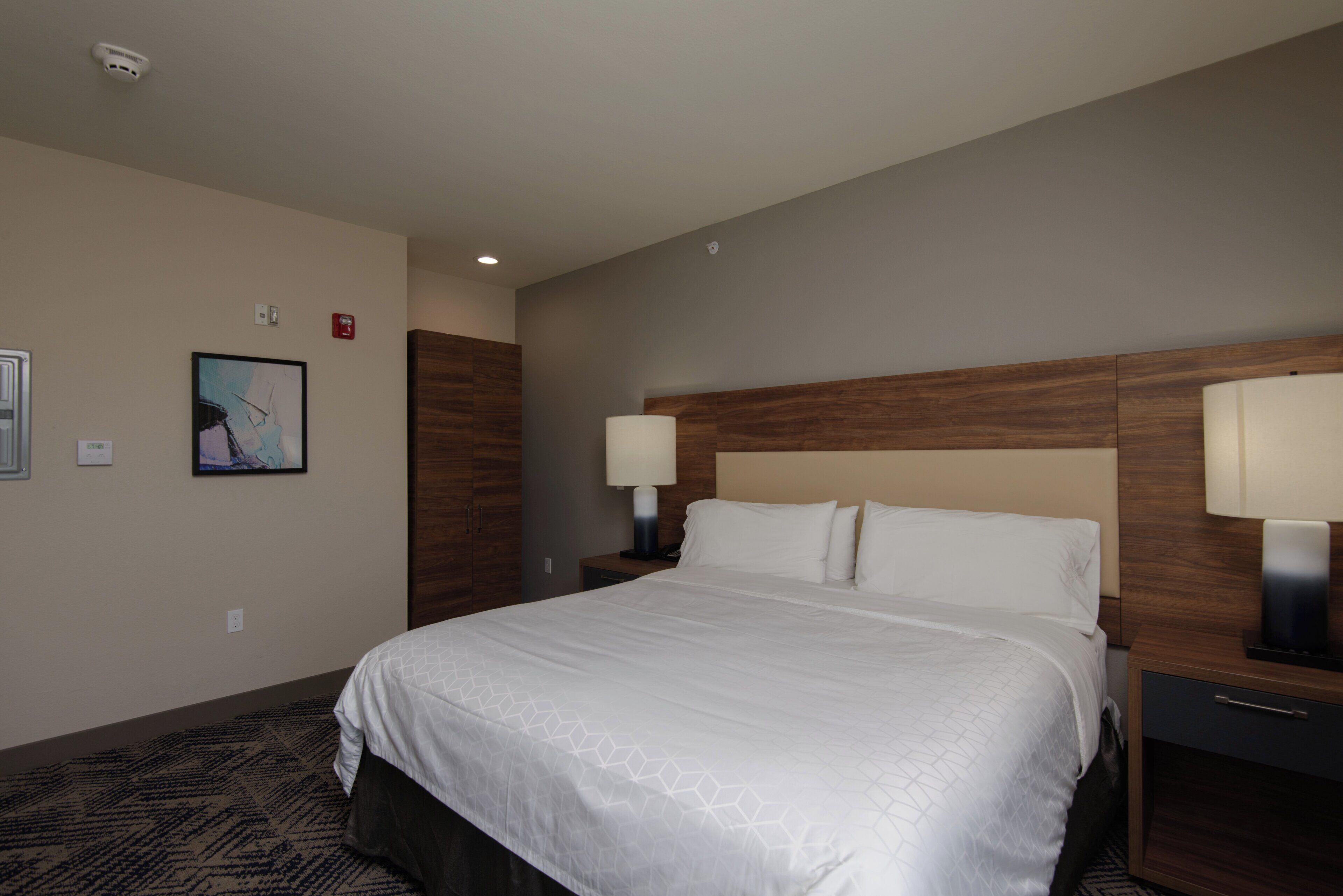 A hotel room with a large bed, two bedside tables with lamps, a headboard, a painting on the wall, and a carpeted floor.