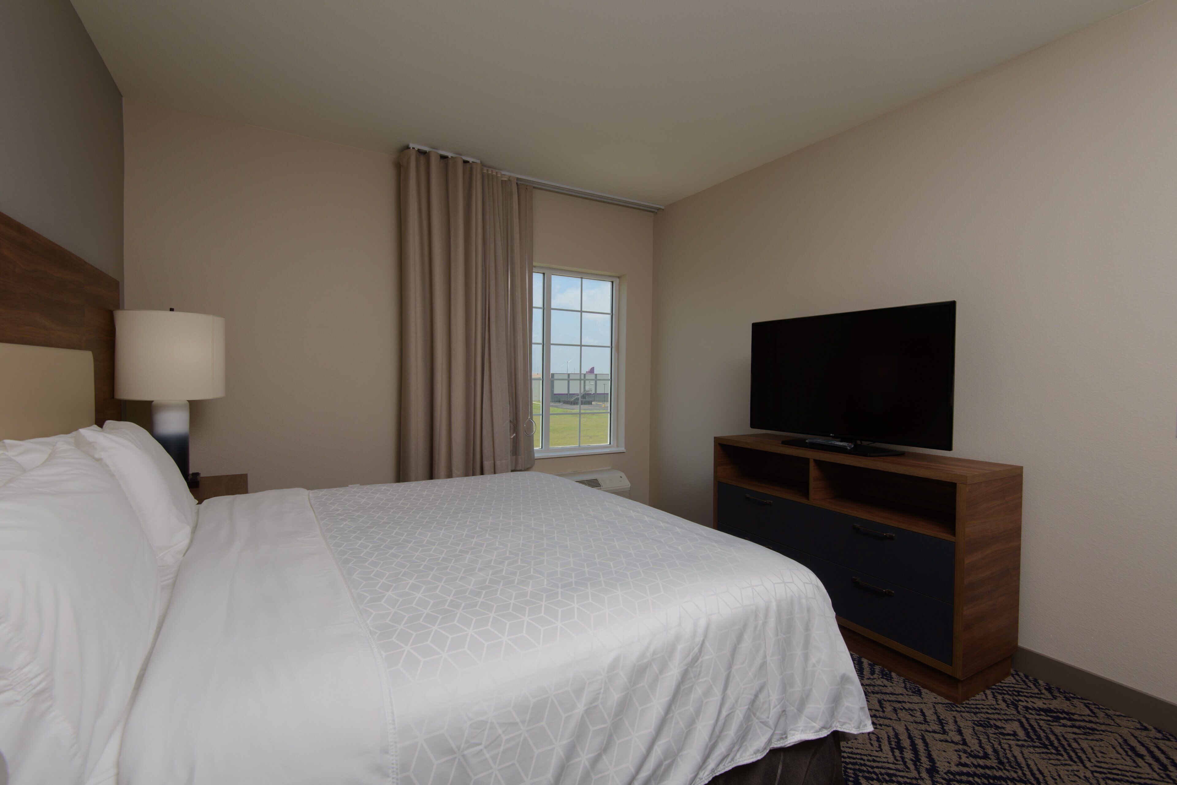 Una habitación de hotel con una cama, un televisor sobre un soporte de madera, una ventana con cortinas y una lámpara en una mesita de noche.