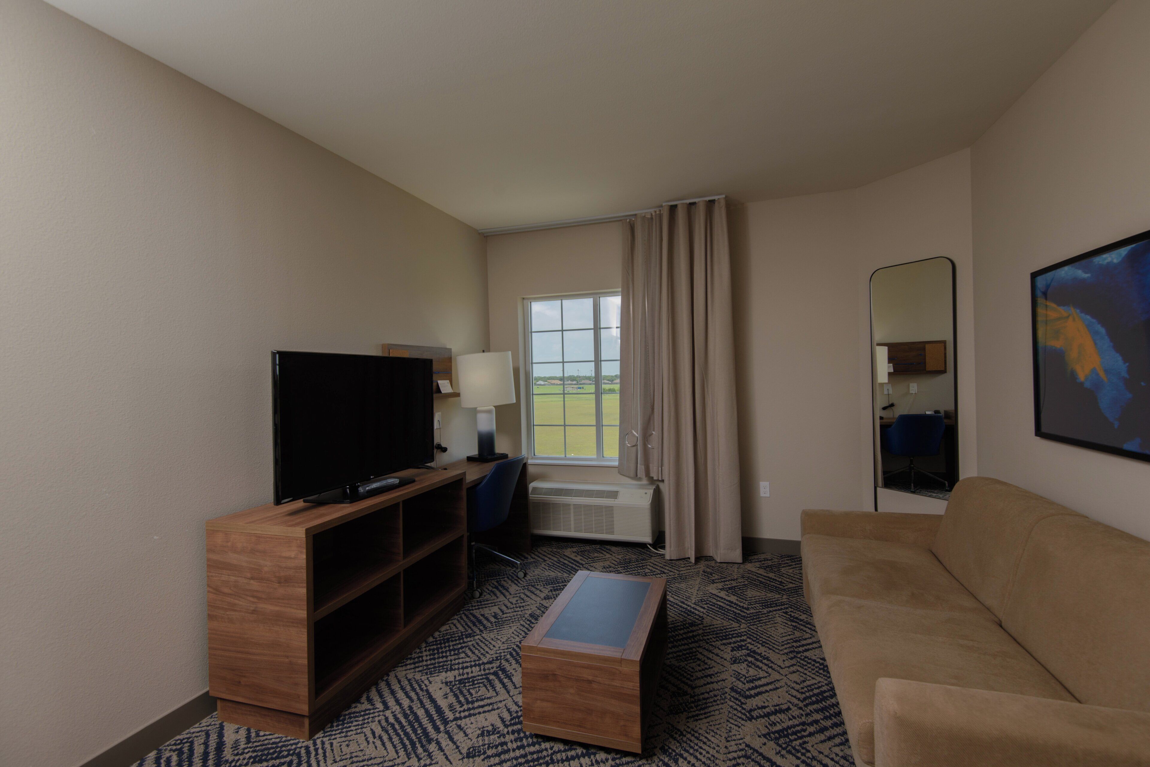A hotel room with a television, a desk, a chair, a sofa, a window with curtains, and a painting on the wall.