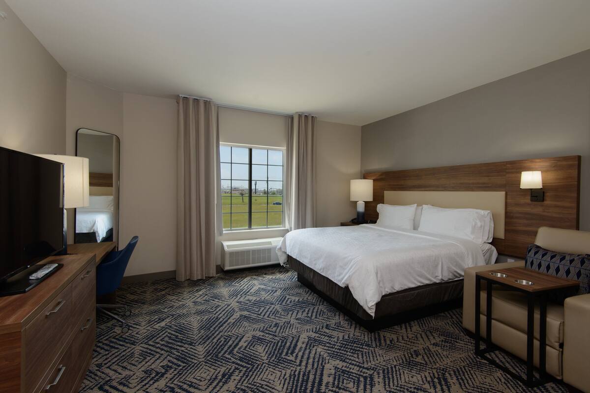 A hotel room with a large bed, a desk with a TV, a chair, a small table, and a window with curtains.