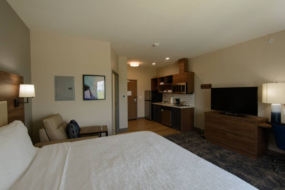 A hotel room with a large bed, a desk, a chair, a television, a kitchenette, and a bathroom.