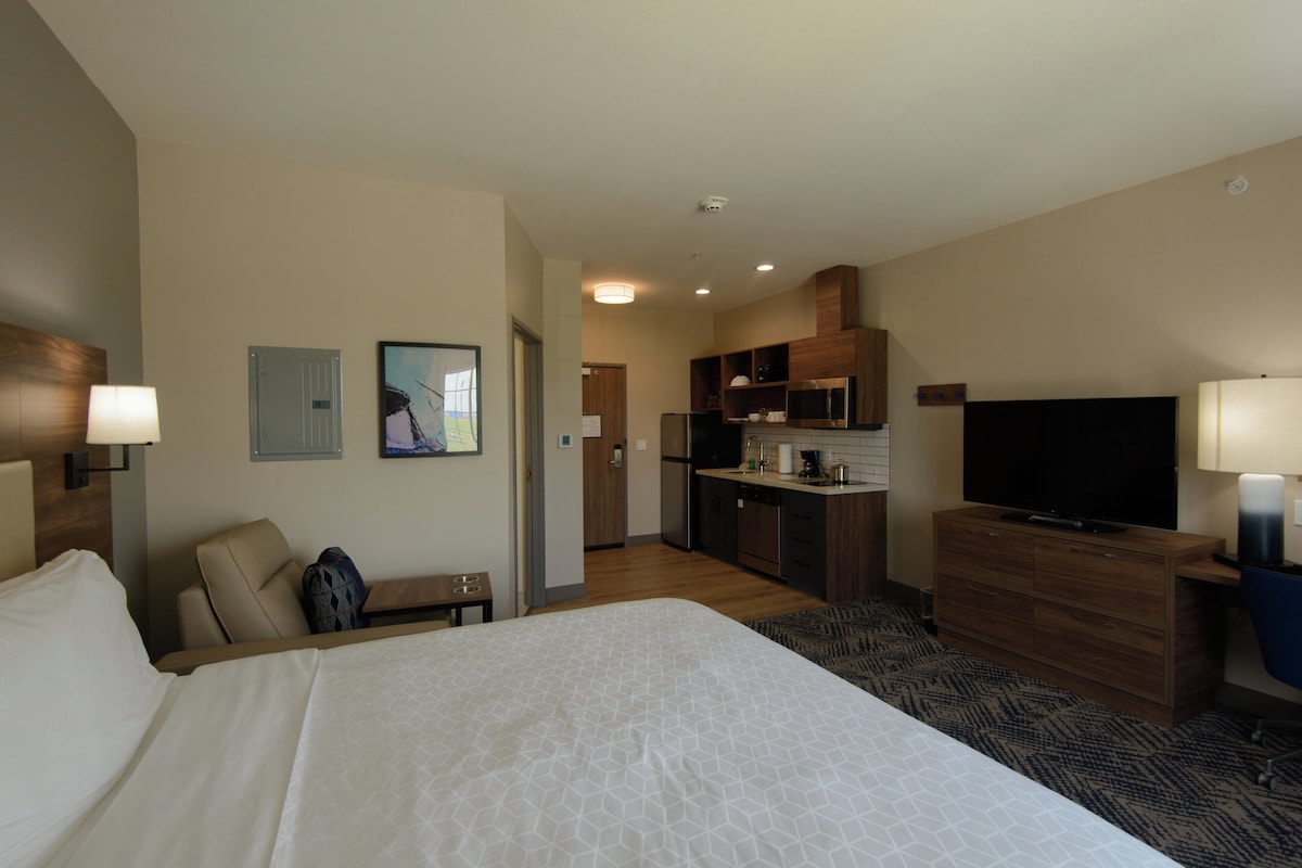 Quarto de hotel com uma cama grande, uma mesa, uma cadeira, televisão, cozinha compacta e banheiro.