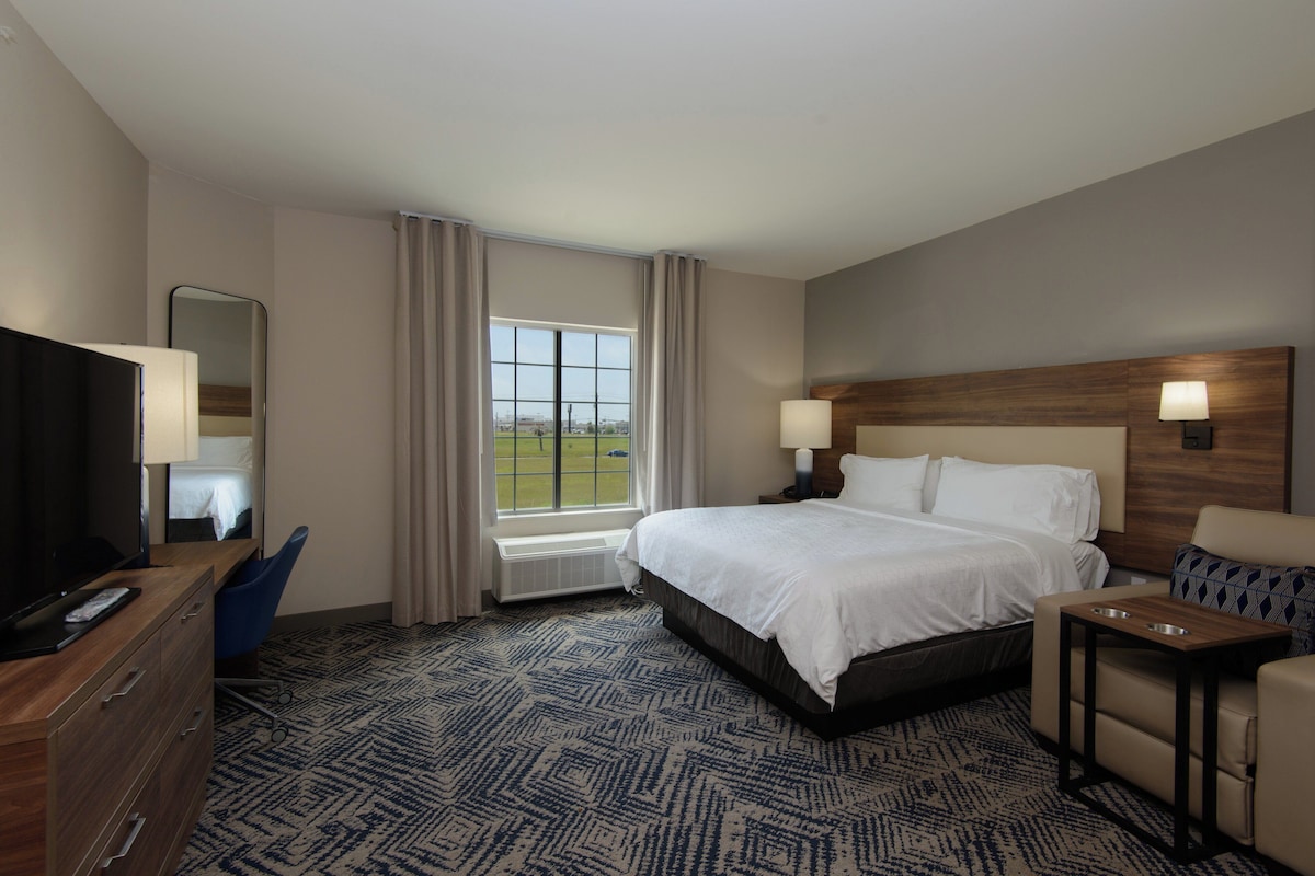 Quarto de hotel com uma cama grande, uma mesa com televisão, uma cadeira, uma pequena mesa e uma janela com cortinas.