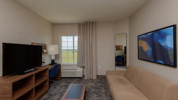 A hotel room with a TV, a sofa, a coffee table, a chair, a window with curtains, and a painting on the wall.