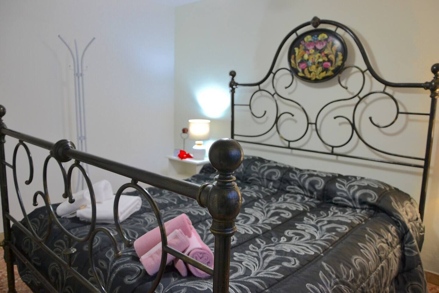Uma cama com cabeceira de metal e colcha estampada, um abajur sobre uma mesa de cabeceira e uma parede com um prato decorativo.