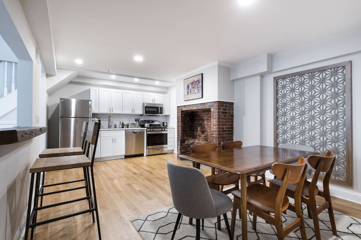 A modern kitchen with white cabinets, a stainless steel refrigerator, a brick fireplace, and a dining area with wooden chairs and a table.
