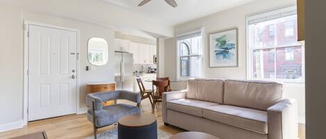 Amplia sala de estar con un sofá color beige, un sillón gris y una mesa de centro de madera. Un ventilador de techo y un ventanal grande ofrecen luz natural.