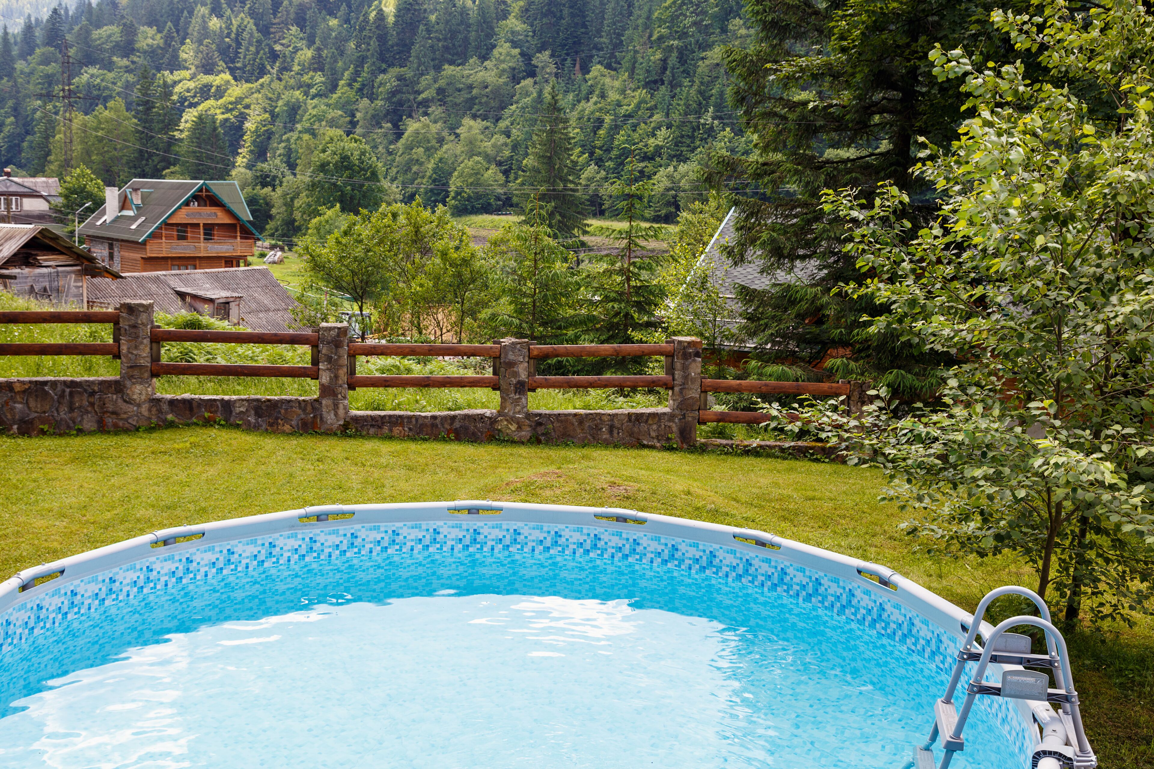 Una piscina con cerca de madera, rodeada de césped y con vistas a casas al fondo.