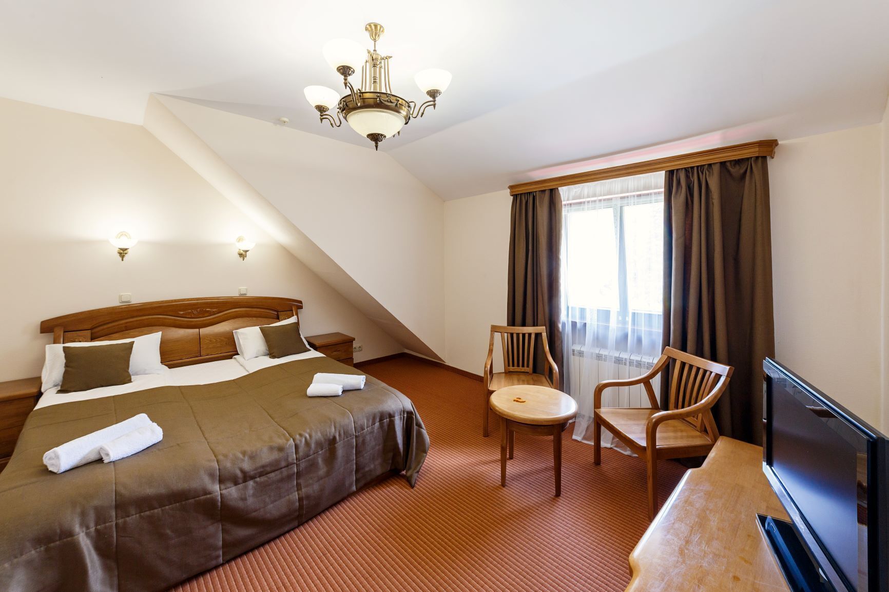 A hotel room with a bed, a desk, two chairs, a television, and a large window with curtains.