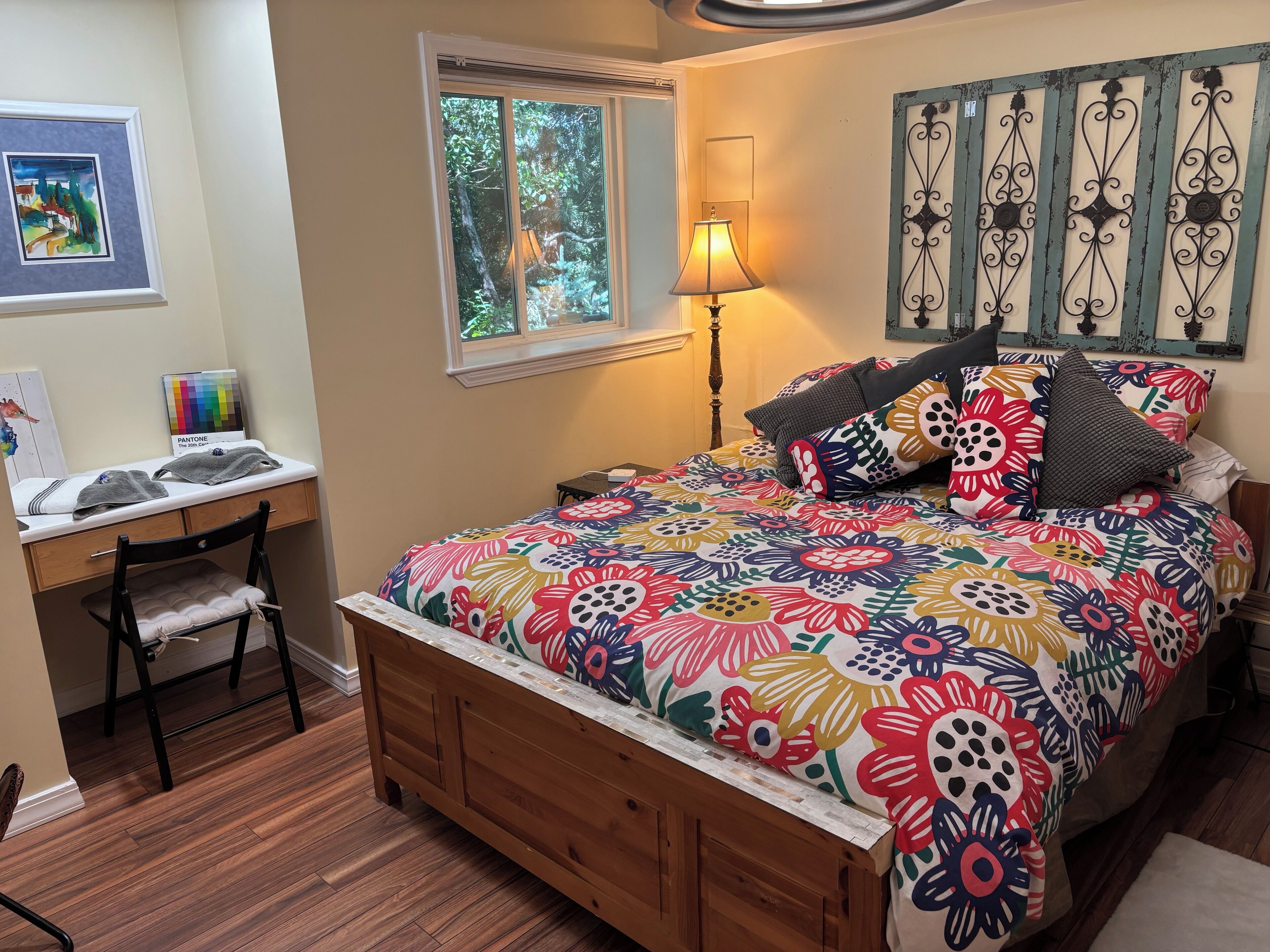 A bedroom with a wooden bed, colorful floral bedding, a desk with a chair, a window with a view of trees, and a wall-mounted decorative piece.