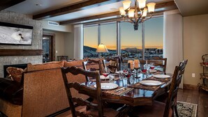 Ein Esszimmer mit einem großen Tisch, gedeckt für sechs Personen, umgeben von Holzstühlen mit gemusterten Kissen. Eine Chandelier hängt darüber, und ein großes Fenster bietet Blick auf eine gebirgige Landschaft.
