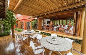 Ein Außenbereich mit einer Terrasse, runden Tischen und Stühlen, einem Holzdeck und einer überdachten Sitzecke mit Kissen und einer Couch.