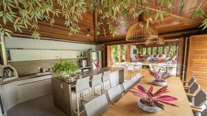 A modern kitchen with a central island, dining area with chairs, and hanging plants. - Spectacular Luxury Mansion on the Beach. (Mata de São João)