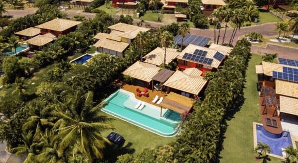 Eine Vogelperspektive zeigt einen Urlaubsort mit mehreren Bungalows, einem zentralen Schwimmbecken und umliegenden Palmen.