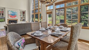 Un coin repas avec une table en bois dressée pour quatre personnes, entourée de chaises en osier, et offrant une vue sur un paysage verdoyant grâce à de grandes fenêtres.