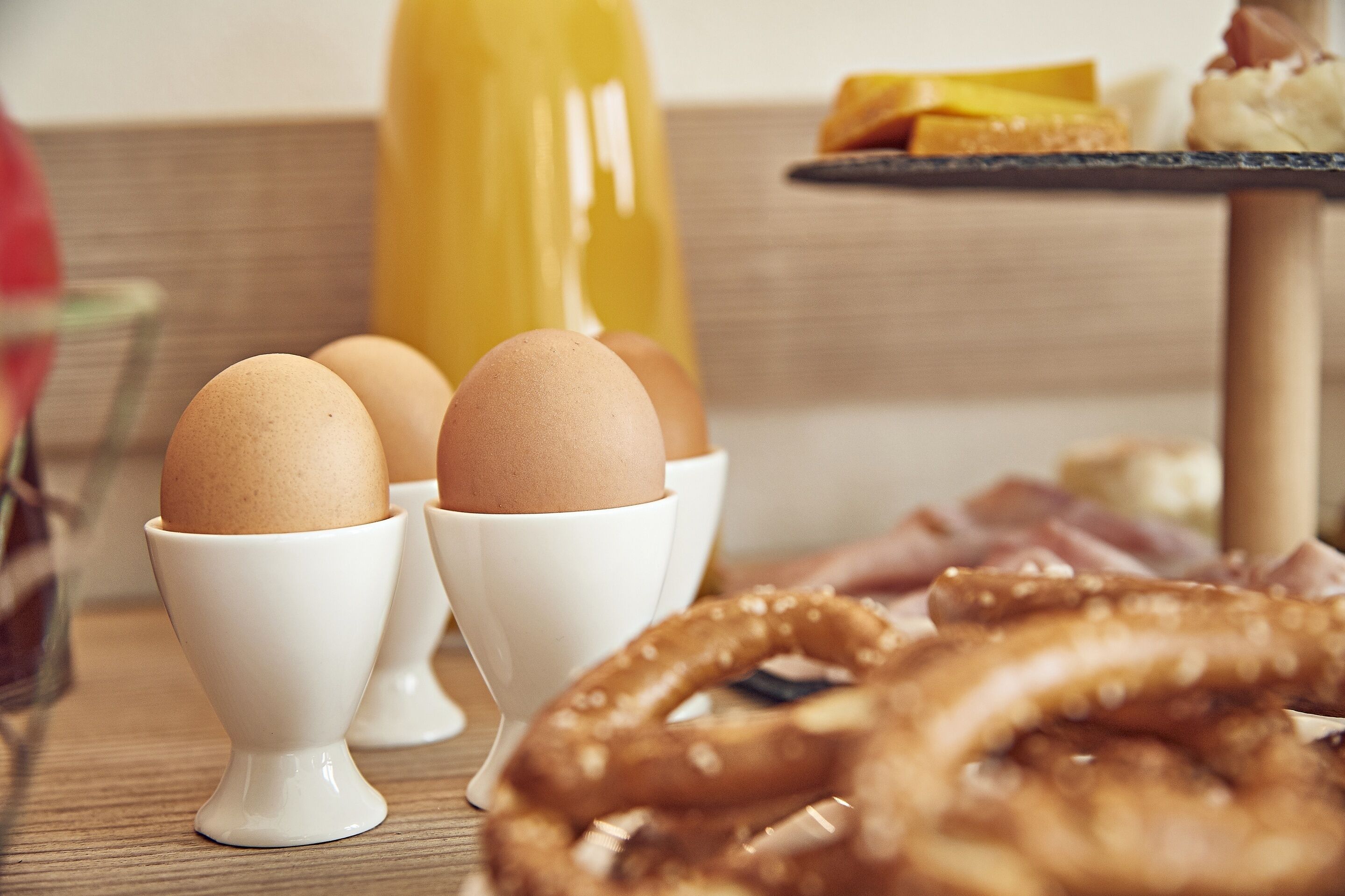Huevos en tazas blancas sobre una mesa con palitos de pretzel y una botella de jugo.
