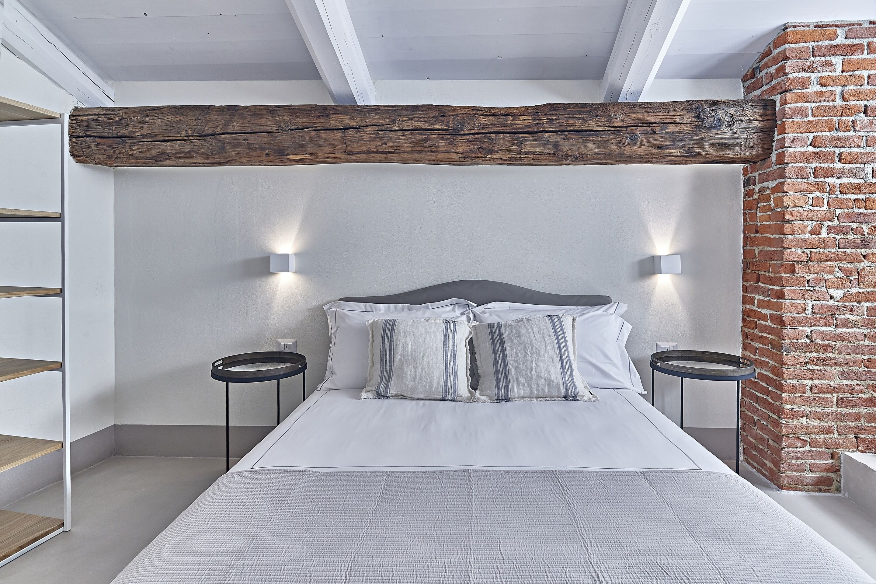 A bedroom with a large bed, white bedding, two bedside tables, a brick wall, and exposed wooden beams.