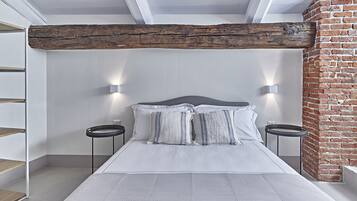 A bedroom with a large bed, white bedding, two bedside tables, a brick wall, and exposed wooden beams.