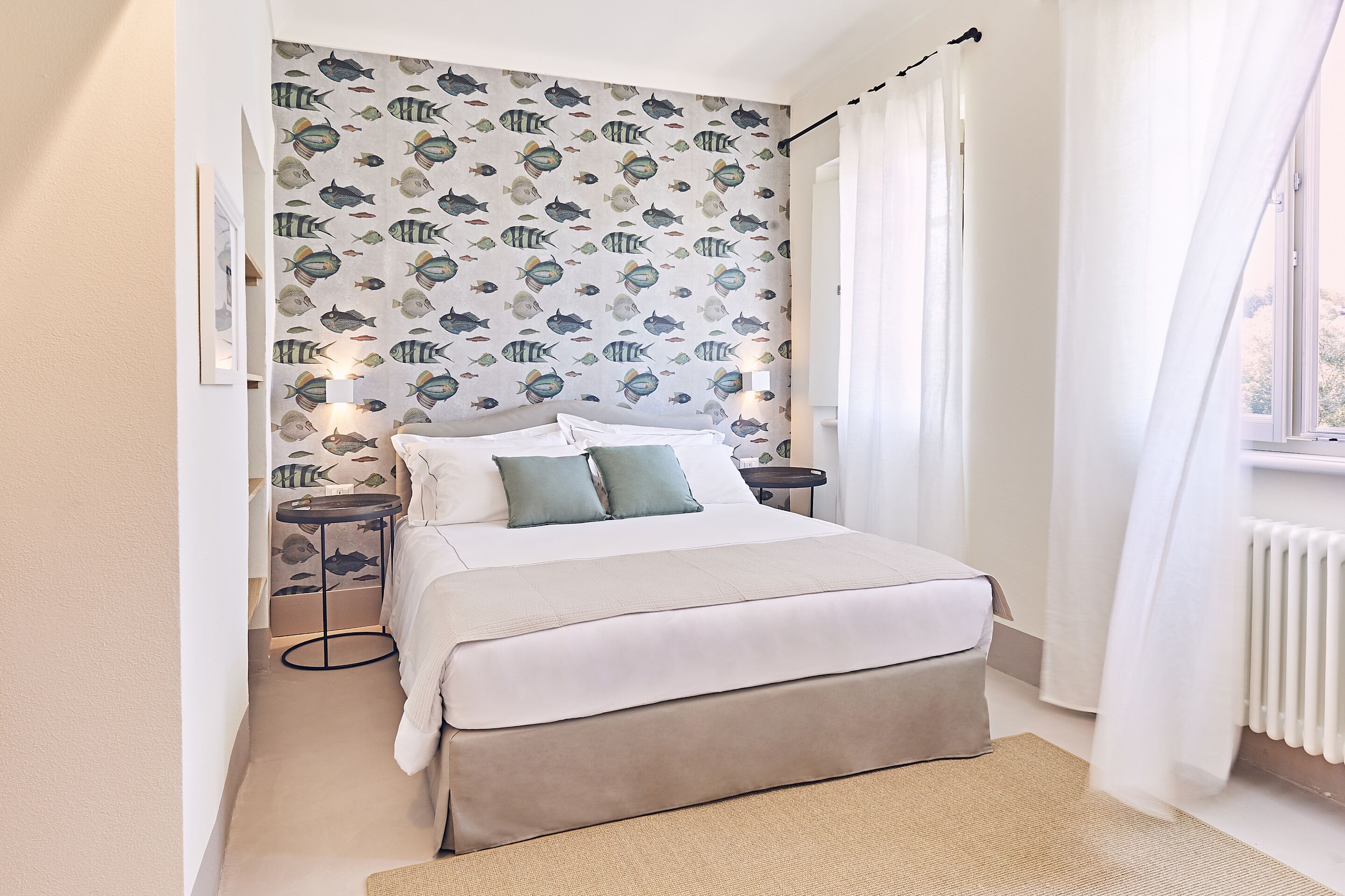A neatly made bed with a white and beige duvet, a small bedside table with a lamp, and a patterned wallpaper with fish designs.