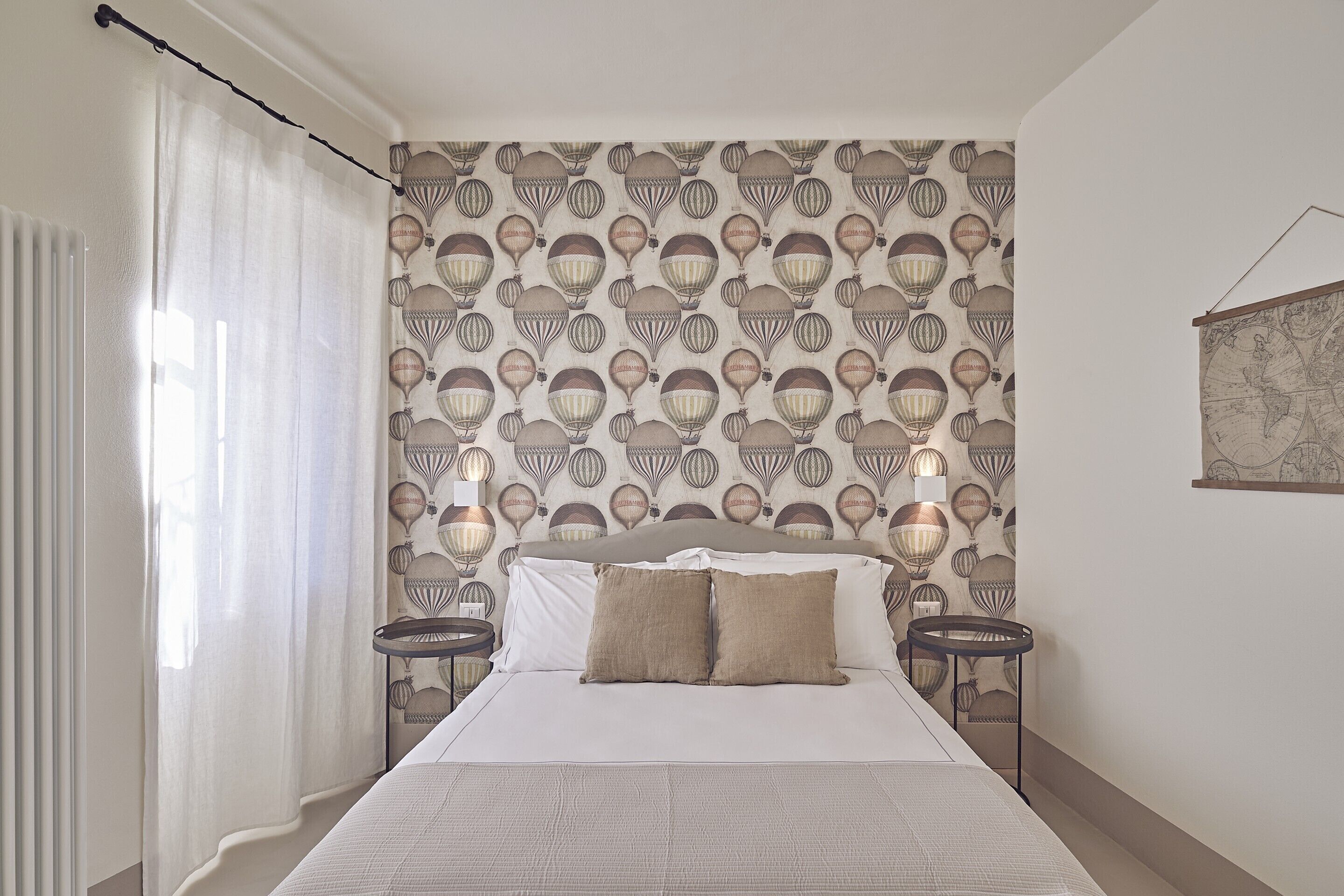 Una cama bien hecha con sábanas blancas y dos almohadas color beige, una pared con un estampado de globos aerostáticos, y dos mesitas de noche con lámparas.
