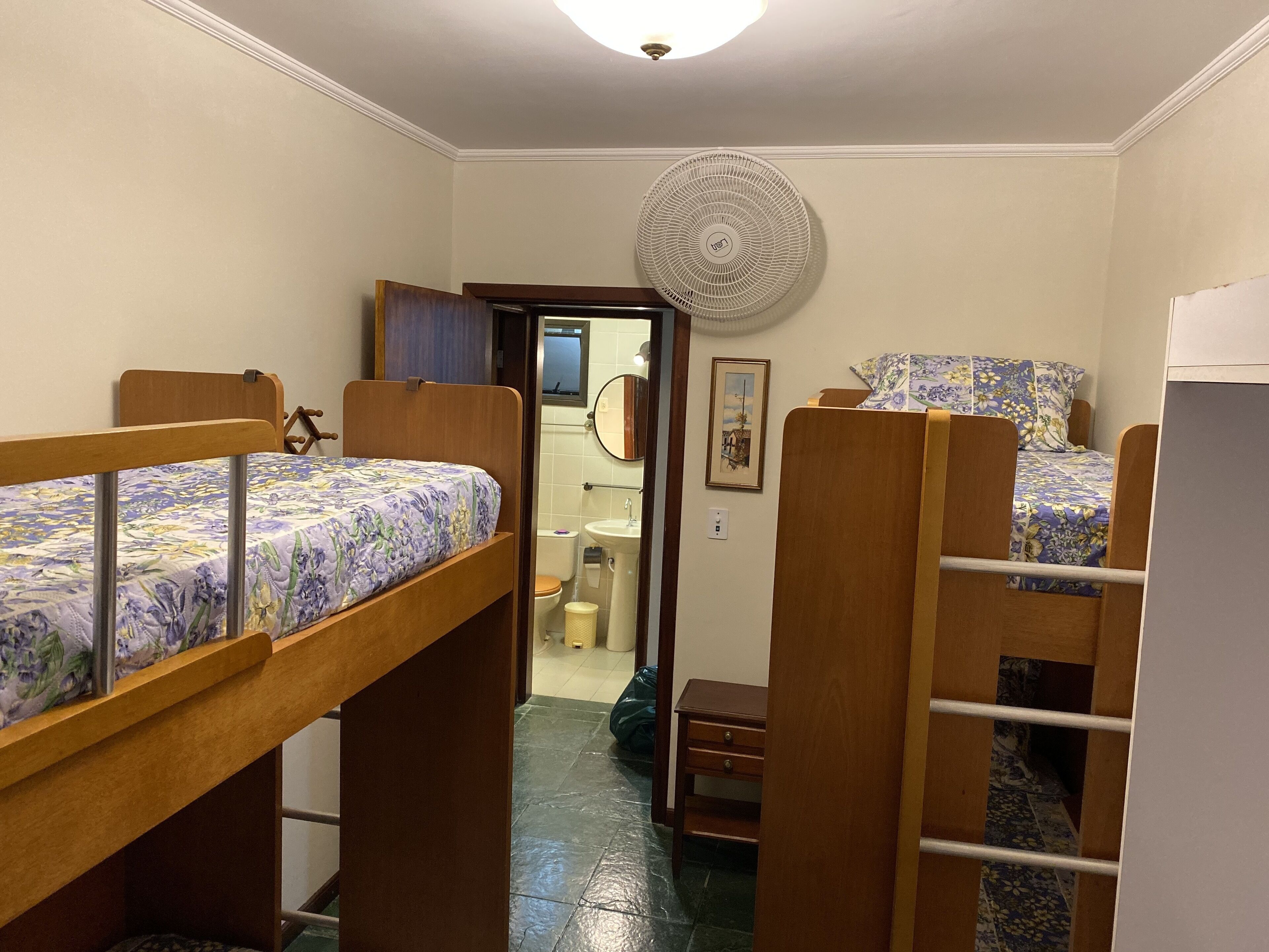 Une chambre de taille modeste, équipée de lits superposés, d’une petite table de chevet, d’un miroir et d’un ventilateur fixé au mur.