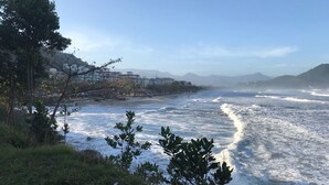 A coastal view with waves crashing onto the shore, a grassy area with trees, and buildings in the background. - Apartment in Praia Grande 100 m from the beach (Ubatuba)
