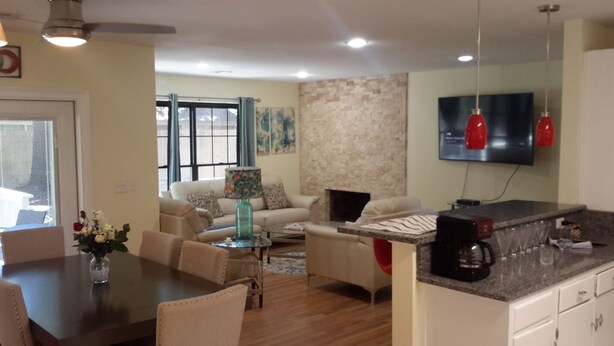 A modern living room with a sofa, coffee table, and dining area. There is a fireplace, a TV mounted on the wall, and a kitchen counter with a coffee maker. - COOL HAVEN- A Quiet, Elegant, Contemporary, Incognito Retreat near FAMU/FSU (Tallahassee)