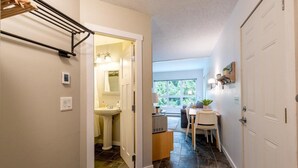 A hallway leading to a bathroom with a sink, a toilet, and a shower. There is a small table with a chair and a plant. The floor is tiled, and there are light fixtures on the ceiling.