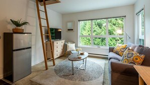A compact living space with a kitchenette, a sofa, a coffee table, and a large window offering a view of greenery.