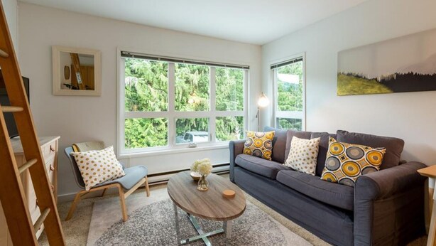 A living room with a grey sofa, a wooden coffee table, and a patterned armchair. There are large windows with a view of greenery outside.
