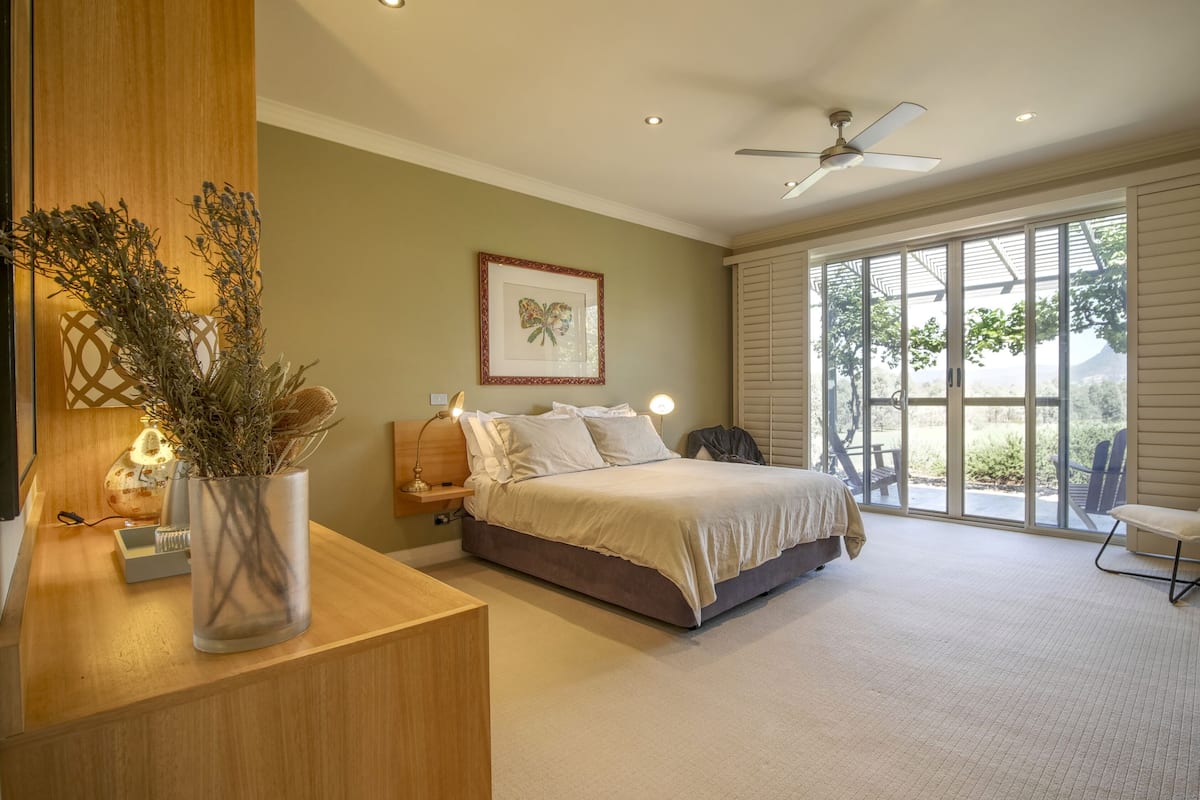 A bedroom with a bed, a bedside table with a vase, a framed picture on the wall, and a large window with a view of greenery.