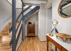 A wooden staircase with a dark railing, a wooden cabinet with a bowl and a bottle, and a round mirror on a white countertop. - George Bass Drive 569 On The Beach Living (Malua Bay)