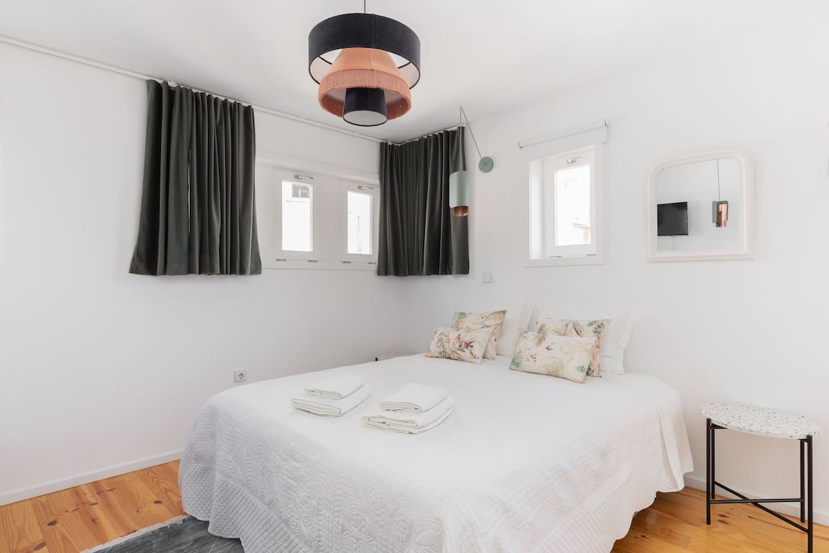A bedroom with a bed, two windows, a hanging light fixture, a small table, and a mirror on the wall.