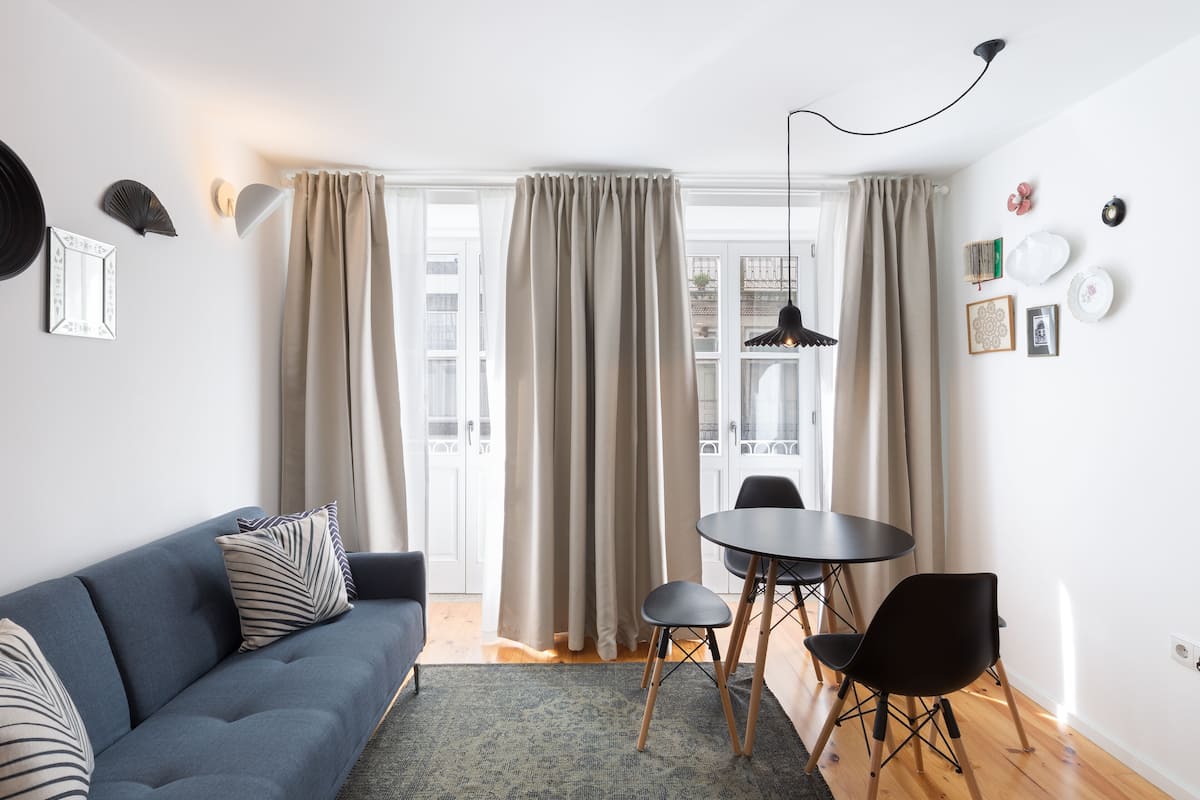 A modern living room with a blue sofa, a round table with chairs, and a large window with curtains.