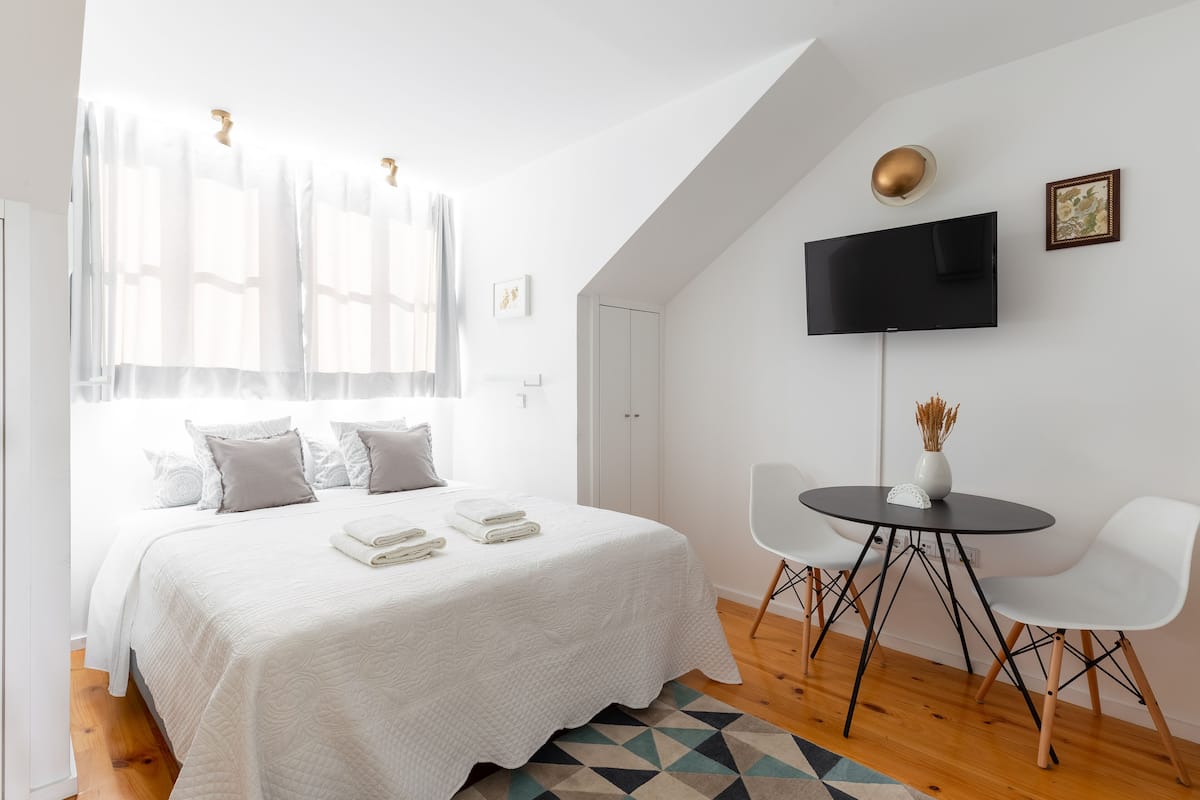 A bedroom with a bed, a TV, a small table with chairs, and a window with curtains.