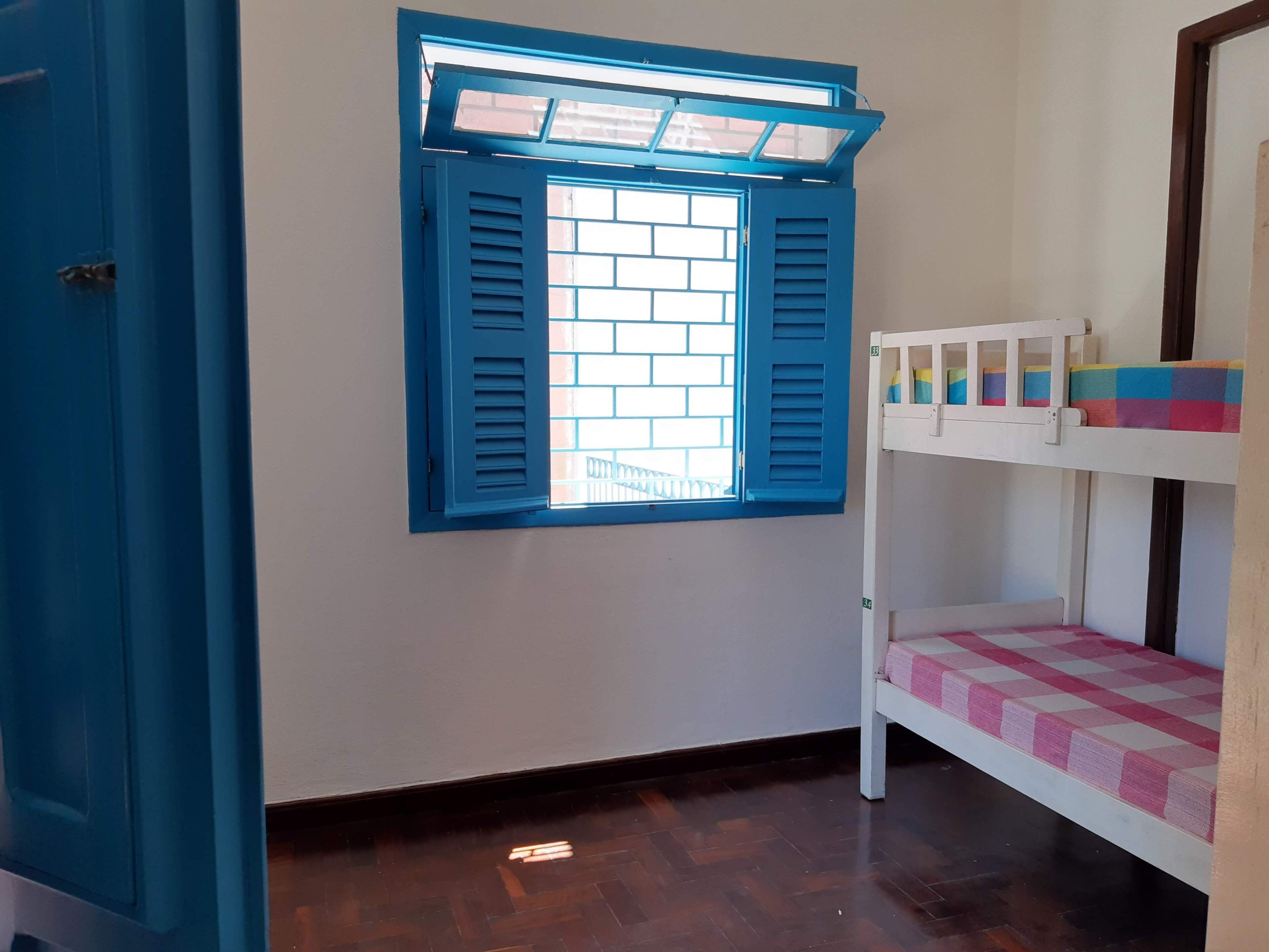A room with a window with blue shutters, a bunk bed with a pink and white checkered bedspread, and a white door.